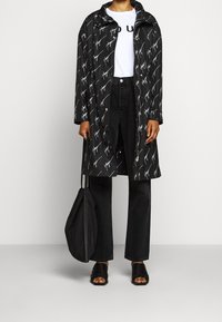 Osoba ubrana w czarny wzorzysty długi płaszcz na białym t-shircie z grafiką i czarnych szerokich dżinsach, trzymająca czarną torbę na ramię i nosząca czarne sandały na obcasach.