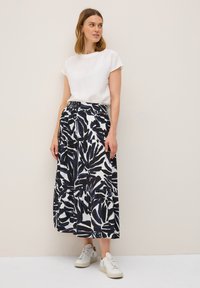 Haut en coton blanc à manches courtes associé à une jupe imprimée longueur genoux avec des motifs de feuilles noires et marines, portée avec des baskets blanches.