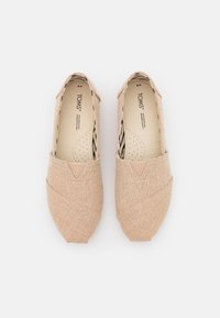Chaussures slip-on en toile beige clair avec une finition texturée, présentant un design croisé en tissu et un panneau élastique pour un enfilage facile.