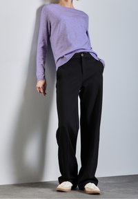 Person wearing a lilac long-sleeve top, black wide-leg pants, and white sneakers standing against a plain light wall.