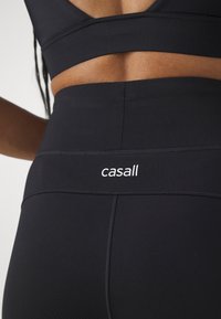 Leggings de sport noirs avec une large ceinture ornée du logo blanc "casall", fabriqués en tissu lisse et extensible. Détails de couture visibles.