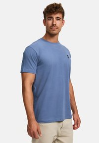 Helles blaues Baumwoll-T-Shirt mit kurzen Ärmeln, rundem Halsausschnitt und einem kleinen schwarzen Logo auf der Brust. Glatte Textur und lässiger Schnitt.