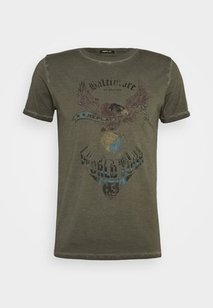 Camiseta de manga corta de color verde oliva con puños enrollados, que muestra un gráfico desvanecido de un águila y un globo terráqueo, con el texto "Baltimore World Tour 85" en el frente.