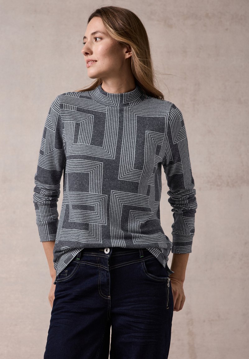 Maglietta grigia a maniche lunghe con un motivo geometrico a linee bianche, colletto a dolcevita a costine e vestibilità morbida, abbinata a pantaloni in denim scuro.