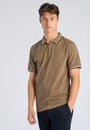 Ung mand iført brun kortærmet poloshirt med lynlås og marineblå bukser, stående mod en ensartet lysegrå baggrund.