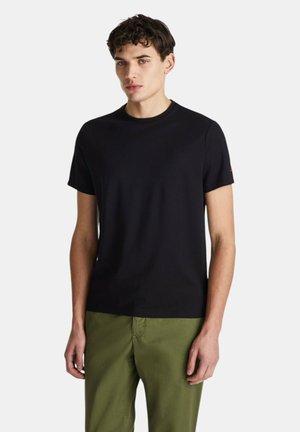 Giovane uomo in piedi con una maglietta nera a maniche corte tinta unita e pantaloni verde oliva su sfondo bianco.