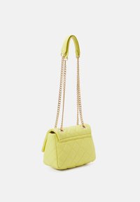 Liten gul quiltad handväska med guldkedja och gul läderaxelplatta som hänger mot vit bakgrund.