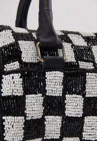 Sac à main en perles noir et blanc avec un motif à carreaux et une sangle en cuir noir attachée par un anneau métallique.