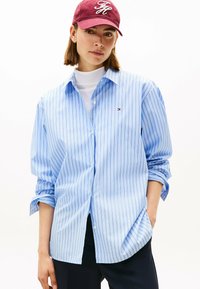 Camicia a righe blu e bianche con maniche arrotolate e vestibilità ampia, con un piccolo logo sul petto. Cappellino borgogna con logo.