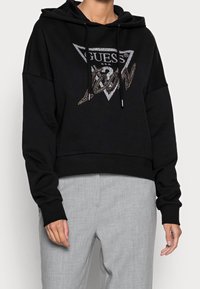 Black cropped hoodie made of soft material featuring a triangle logo "GUESS" in silver and a textured gold accent. Drawstring hood included.