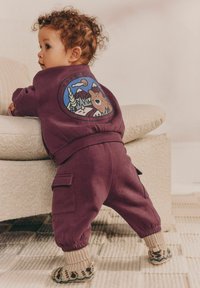 Lilla sweatshirt og bukser sæt til småbørn, med et rundt bjørne grafik på ryggen. Teksturede sokker med et mønstret design fuldender outfittet.