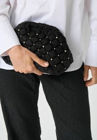 Mustast pehmest materjalist kokku õmmeldud käekott, millel on kogutud ülemine osa ja millel on väiksed hõbedased detaillid, hoitakse käes.