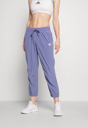 Kvinde iført hvid sports-bh, lilafarvede Adidas joggebukser og hvide sneakers, stående mod en ensfarvet lys baggrund.