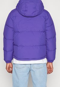 Fialová prošívaná bunda s kapucí, na zádech je vyšité logo Lacoste. Bunda má prošívaný design a elastické manžety.