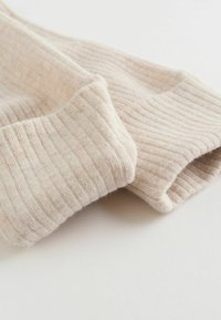 Zbliżenie na beżowe rękawy z prążkowanego dzianinowego materiału z podwiniętymi mankietami na jasnej powierzchni.