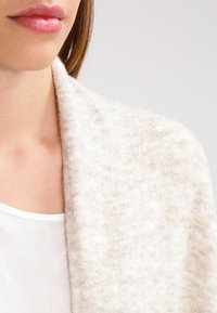 Helle beige gestrickte Schal mit weicher Textur, mit einem breiten Kragen und subtilen Farbvariationen, der über einem weißen Kleidungsstück liegt.