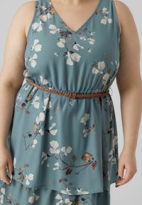Blomsterkjole i teal stof med V-udskæring, lagdelt design og et flettet brunt bælte, der fremhæver taljen. Hvide og brune blomster mønstre.