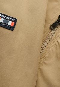 Tissu beige avec une texture à fines côtes, présentant un patch Superdry bleu marine avec des accents rouges, blancs et noirs, ainsi qu'un détail de fermeture éclair.