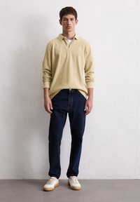 Jasnożółta, długosleeve'owa koszulka polo, ciemnoniebieskie dżinsy i białe sneakersy. Luźny krój z zapięciem przy kołnierzyku. Gładka faktura i czysty design.