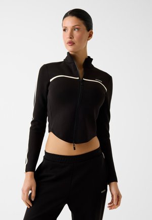 Moteris dėvi juodą trumpą striukę su baltomis detalėmis ir derančias juodas kelnes, pozuoja prieš balto fono foną.