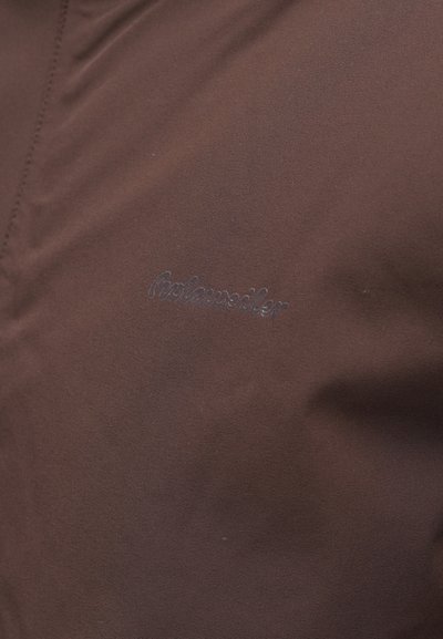 Brūns audums ar gludu tekstūru. Ar šuvju detaļām un smalku embosētu logotipu tumšākā brūnā krāsā.