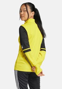 Gelbe Sportjacke mit schwarzen Ärmeln, hohem Kragen und gestreiften Akzenten. Entwickelt für Komfort und Beweglichkeit. Glatte Textur.