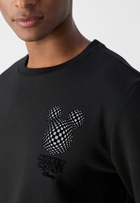 Sudadera negra con un diseño texturizado de Mickey Mouse en forma de cuadro en el lado izquierdo del pecho, con texto bordado "Mickey Mouse" debajo.