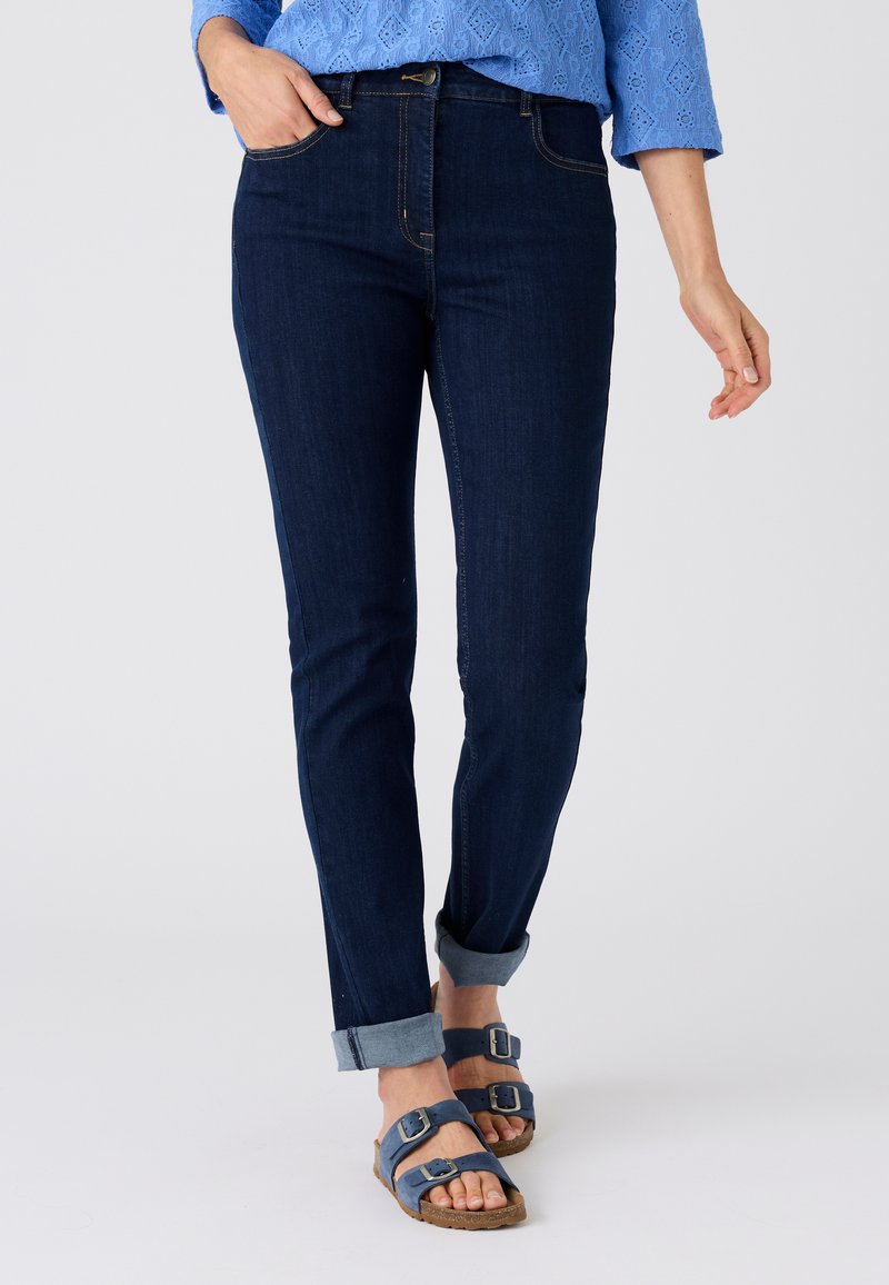 Frau trägt dunkle, hochgekrempelte Skinny-Jeans, blaue Sandalen mit Schnalle und ein blau gemustertes Oberteil, steht vor einem einfarbigen Hintergrund.