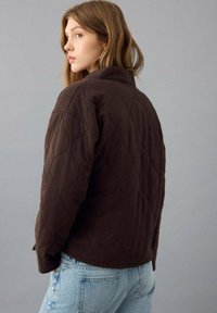 Braune gesteppte Jacke mit hohem Kragen und strukturiertem Wellenmuster, kombiniert mit hellblauen Jeans. Blick von hinten.