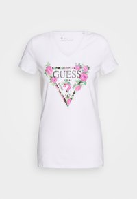 Camiseta blanca de cuello en V con un gráfico de triángulo floral que presenta la palabra "GUESS" adornada con strass y acentos de hojas verdes.