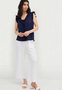 Marineblauw gerimpeld tanktop met een V-hals en knoopdetails aan de voorkant, gecombineerd met losse witte linnenbroek en crèmekleurige blokhak-sandalen.