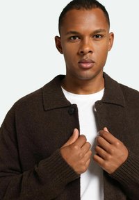 Cárdigan marrón de punto con cuello de solapa, cierre de botones y puños acanalados. Lleva puesto sobre una camisa blanca, mostrando manos agarrando la tela.