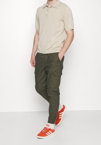 Beige Poloshirt mit kurzen Ärmeln und Kragen, olivgrüne Cargo-Hosen mit Seitentaschen und orangefarbene Wildleder-Sneaker mit weißen Sohlen.