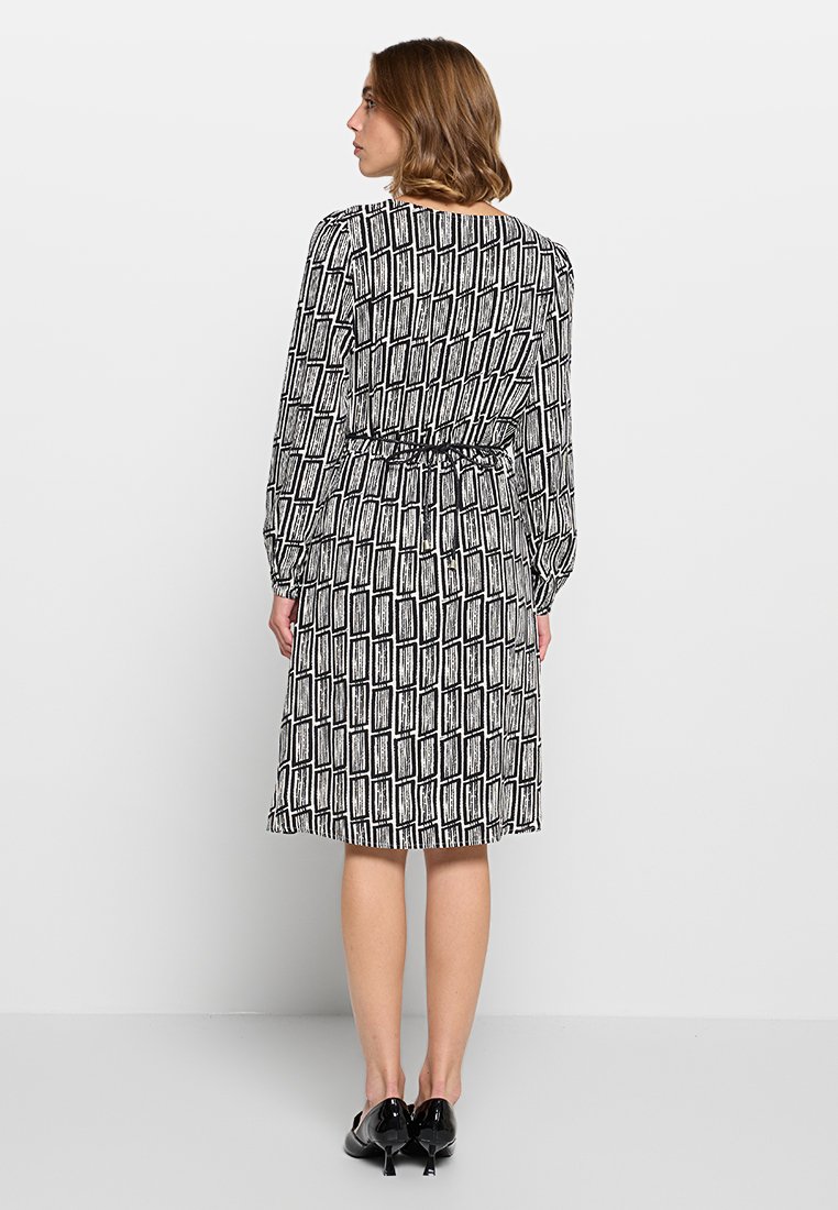 Vestido de patrón en blanco y negro con mangas largas, cintura ceñida y dobladillo a la altura de la rodilla. Tela suave con diseños rectangulares.