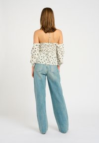 Donna di spalle che indossa un top bianco floreale con spalle scoperte e maniche lunghe, abbinato a jeans larghi di colore azzurro chiaro, su uno sfondo semplice.