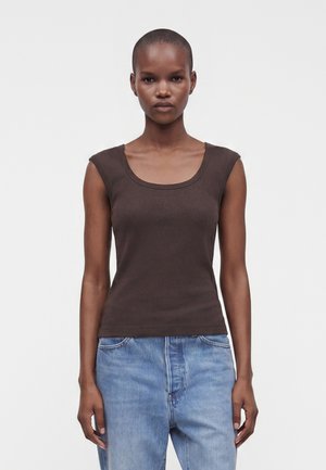 Femme aux cheveux courts portant un haut marron foncé sans manches et un jean bleu clair, debout devant un fond clair uni.