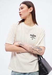Krémovo sfarbené bavlnené tričko s krátkymi rukávmi, s čiernym vyšívaným textom "THE DANCE STUDIO" a úzkym strihom. Doplnkom je tmavá taška.