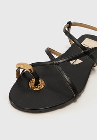 Sandal preto plano com tiras de pele cruzadas, forma arredondada, detalhe circular em ouro com texturização e sola lisa.
