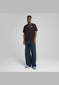 Sort bomulds T-shirt med et brystgrafik, parret med løse mørke denimjeans og blå sko med en brun sål.