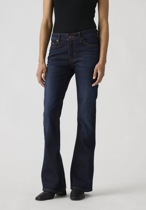 Kobieta w ciemnoniebieskich jeansach bootcut i czarnym topie bez rękawów, stojąca na jasnoszarym tle.