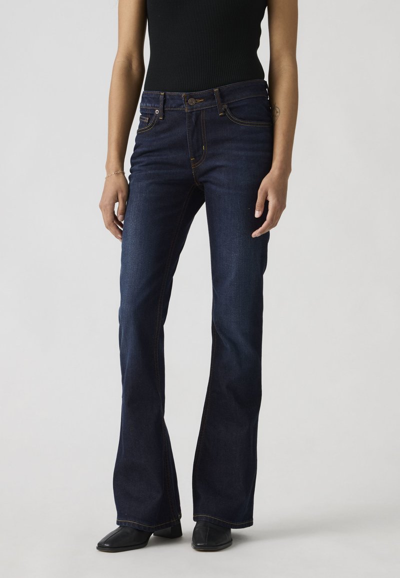 Femme portant un jean bootcut bleu foncé et un top noir sans manches, debout devant un fond gris clair.