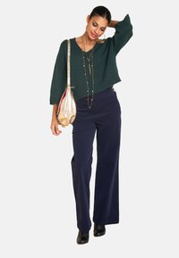Grüner Strickpullover mit weiten Ärmeln, kombiniert mit navyblauen weit geschnittenen Hosen. Eine goldene Crossbody-Tasche und geschichtete Halsketten runden den Look ab. Schwarze Stiefel.