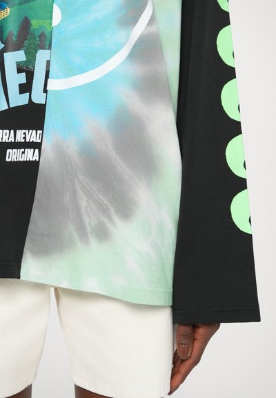 Majica dugih rukava s grafikom koja prikazuje crne, zelene i tie-dye uzorke; opušteni kroj s prostranom rukavom, te tiskanim tekstom i akcentima.