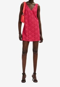 Modelka oblečená v živě růžových květinových krajkových mini šatech, doplněná malou korálovou kabelkou a černými sandály na vysokém podpatku, pózující na bílém pozadí.