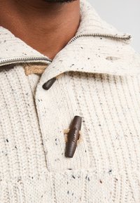 Kerman neulepusero, jossa on korkea kaulus, vetoketju ja puinen solki suljento. Kankaan kautta kulkevat hienot läikät tuovat tekstuuria.