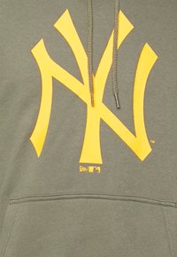 Olivgrön hetsare med en stor gul NY-logotyp, snörning och framficka. Tillverkad av mjukt material med en snug passform.