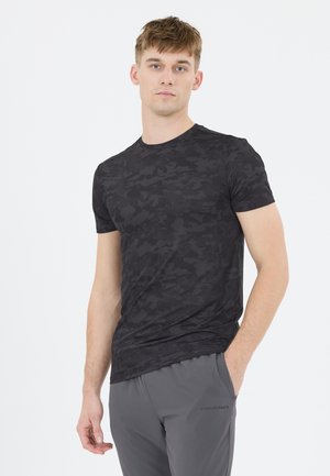 Ein Mann trägt ein eng anliegendes schwarzes Camouflage-T-Shirt und graue Sporthosen und steht vor einem schlichten weißen Hintergrund, nach vorne blickend mit neutralem Gesichtsausdruck.