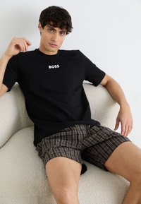 Schwarzes Baumwoll-T-Shirt mit "BOSS"-Logo, kombiniert mit braunen karierten Shorts in Orange- und Gelbtönen, während man auf einem beigen Sofa sitzt.