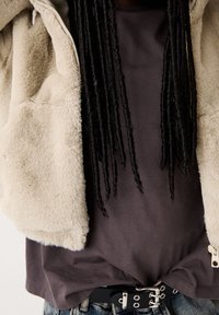 Giacca beige pelosa sopra una camicia grigio scuro, caratterizzata da una cintura nera con accenti in metallo. Materiali strutturati e aspetto stratificato.