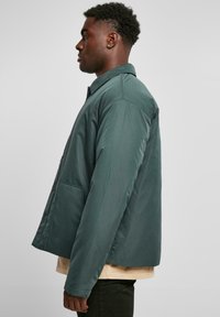 Mørkegrønn puffjakke med krage-design, frontlukking med knapper og sidelommer. Glatt tekstur med en litt oversized passform.